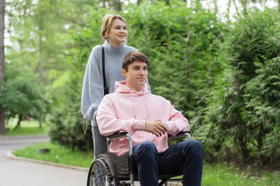 A mother pushes her teenage son in a wheelchair through a park